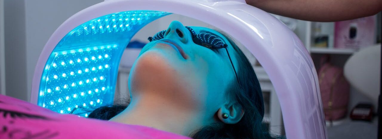 Unrecognizable woman with dark hair with pierced ears lying under ultraviolet lamp while visiting contemporary clinic and spending time in modern salon